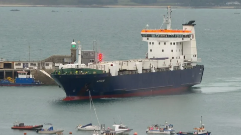 A large red, white and blue vessel travels along calm waters into a harbour area. Smaller boats are moored on buoys in front of the large boat.