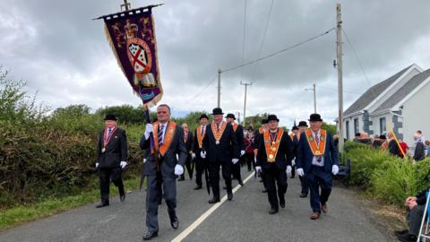 Members of the Orange Order march along a country road, grey skies in the background