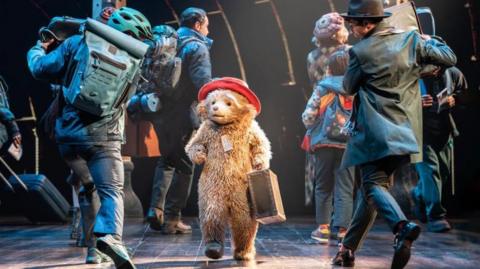 Small bear with red hat and suitcase walks through a busy stage crowd of travellers