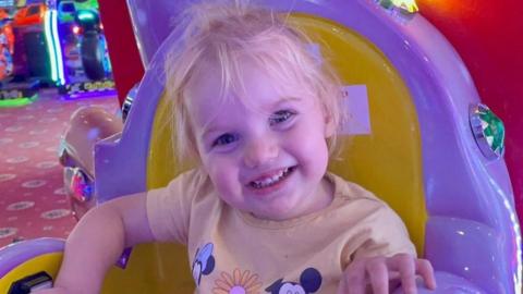 A young blonde toddler wearing a yellow T-shirt smiling into the camera. She is sitting on a yellow and purple chair at an arcade.