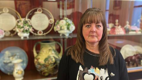 A lady with long brown hair in a black top with a design on and a black cardigan over the top. She is stood in front of various bits of pottery placed on a shelf.