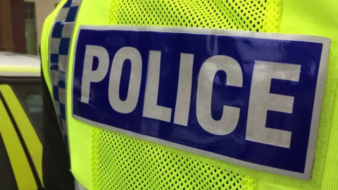 A close-up back view of a police officer, wearing a hi-vis yellow jacket with police written in white letters on a blue background. On the left, a yellow and white police vehicle can just be glimpsed. 