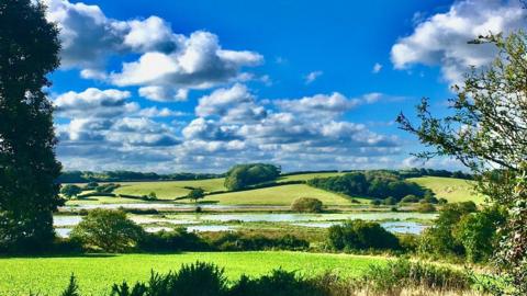 Bright blue sky with white clouds over rolling fields, and some in the centre are flooded.