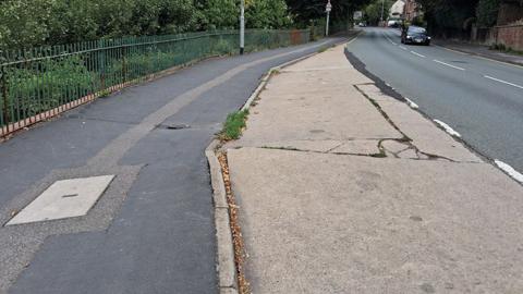 Yarborough Hill in Lincoln a two lane road with green metal fencing on one side and houses on the other the pavement and a layby both show signs of cracking