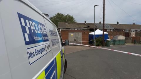 A police investigation scene in a residential neighbourhood.
In the foreground on the left side of the image, a white police van is parked facing away from the camera. The side of the vehicle displays the words "POLICING Yorkshire & The Humber" along with a blue-and-white checkerboard logo. Additional text on the van reads "Crime Scene Investigation" and "Non Emergency 101". Across the middle of the image, red-and-white police tape has been stretched across the road, creating a cordon that restricts access to the area beyond.
In the background, a row of terraced houses stands behind the police cordon. Near the centre of the image, one property shows visible signs of damage, with a large blackened hole in the roof and surrounding scorch marks.
A blue-and-white forensic tent has been erected near the damaged property.