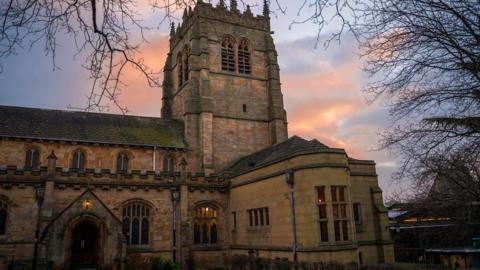 The outside of Bradford Cathedral, pictured against the sky at dusk. A leafless tree stands to the side.