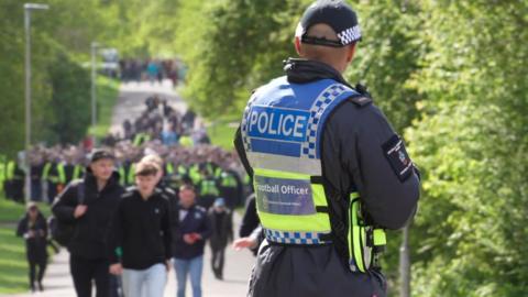 A Devon and Cornwall Police football officer on duty in Central Park near Home Park Stadium in Plymouth. He is in police uniform looking at a large group of supporters and police officers walking towards the ground. There are lots of trees and greenery around him.