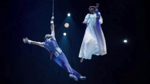 Male acrobat wearing blue hangs from the ceiling while a female acrobat wearing white looks on while hanging 