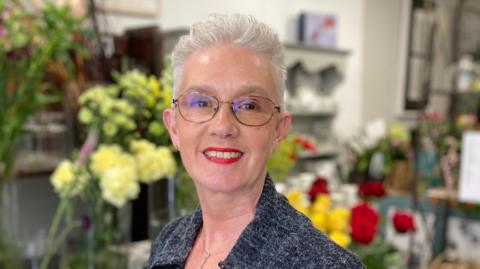 A woman with short grey hair and glasses is wearing a green blouse with grey spots beneath a grey cardigan. She is in front of the blurred images of several flowers.