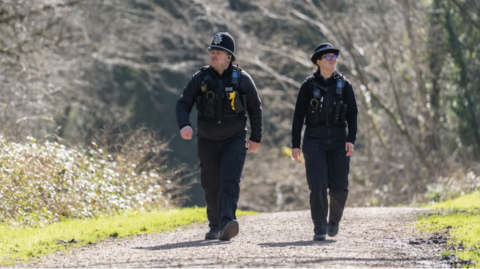 Two police officers, male and female, walking down a footpath. There are trees behind them. One officer is looking to the left and the other is looking to the right.