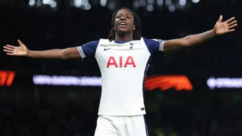 Damola Ajayi stretches his arms out either side in celebration after scoring for Tottenham Hotspur, wearing the club's white home jersey