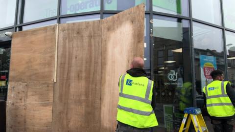 Two workers in high-vis vests taking down wooded boards from a co-op shopfront