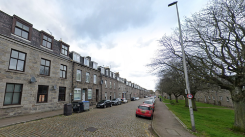 A general view of Jasmine Terrace in Aberdeen. A cobbled street runs through the middle of the image. A red car is parked on the right of the road next to a pavement and a grass verge with bare trees. On the left of the image is a long row of dark-coloured stone flat buildings. The sky is grey with dark clouds in the foreground.