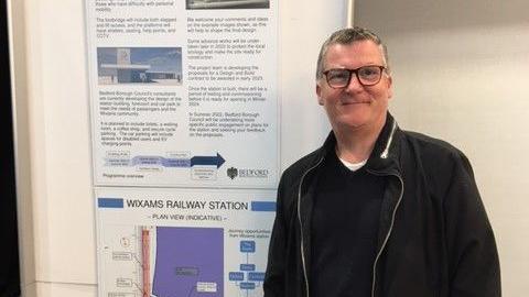 A picture of man in front of a sign with plans for Wixams railway station. He is standing to the left of the sign and is wearing a black jacket with a black jumper underneath. He is wearing brown glasses and has short hair.