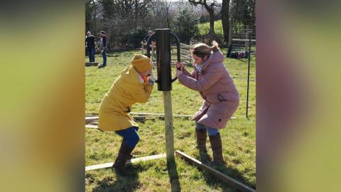 A woman in a yellow coat and hat planting a tree in the ground on farmland along with a woman standing opposite her wearing a pink coat. 