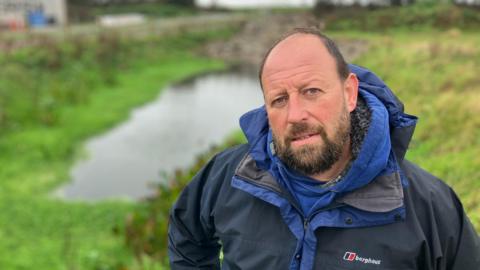 A man stands in front of  a pond on a farm. He is wearing a waterproof coat.   