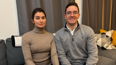 A couple sitting on a grey sofa. William is on the right in a grey jumper whilst Kristina has a light brown turtle neck on, sitting to the left of him. In the background are some cushions along with grey curtains behind them.