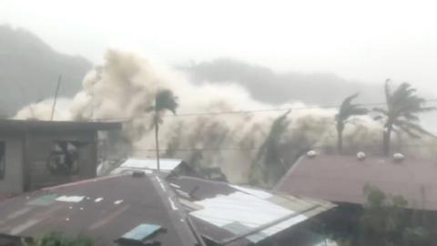 A huge frothy wave crashes over houses and palm trees.