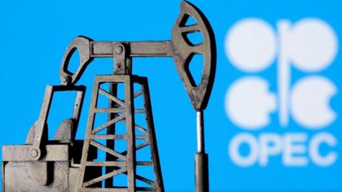 A 3D printed oil pump jack is seen in front of displayed Opec logo in white lettering on a blue background.