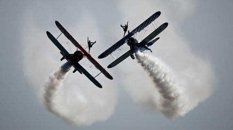 Two planes flying in the sky with smoke trails coming off the back. On the top of each plane is an acrobat.