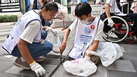 People in Thailand taking part in one of the stages of the Litter Picking World Cup in 2023, a woman and a boy are seen sitting on the ground putting rubbish into a bag