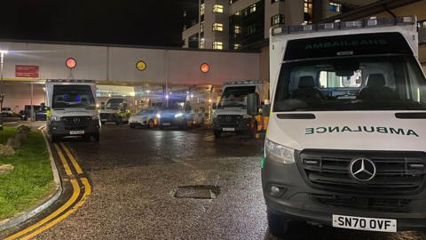 Ambulances with green and yellow checked markings, with lights on, parked outside a hospital building in the dark.