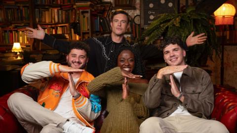 Ed Gamble stands behind a sofa where three former Traitors - Wilfred Webster, Minah Shannon and Harry Clark - sit, making a 'T' sign with their hands