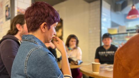 A group of people seated around a wooden table in a casual café setting. The person in the foreground is wearing a denim dress with striped sleeves and has short, dark reddish hair. A paper coffee cup is on the table, and the background shows white tiled walls and a hanging red light fixture.