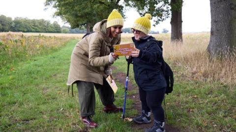 A woman and a girl stand in a field reading a placard. The pair are wearing yellow hats and hiking gear. Trees can be seen behind them.