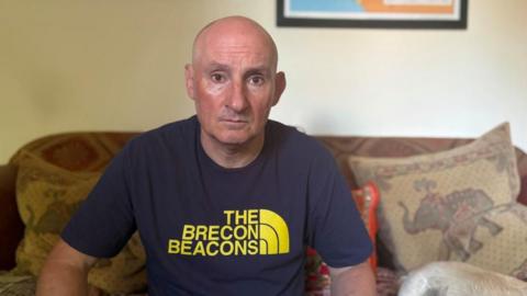 A man with a bald head and a navy T-shirt with yellow writing on it is looking at the camera. He is sitting on a beige sofa with cusions. The wall behind him is magnolia and the base of a framed picture can be seen in the top right corner.