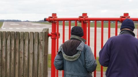 Two people with dark winter coats and hats stand with their backs to the camera in front of a large red metal gate that looks out onto an aircraft runway.