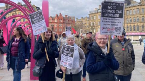 A group of people standing in a square holding and waving banners reading: "No to council tax increase". Behind them is a large pink sculpture and in the distant background are a row of four storey period-looking commercial buildings.