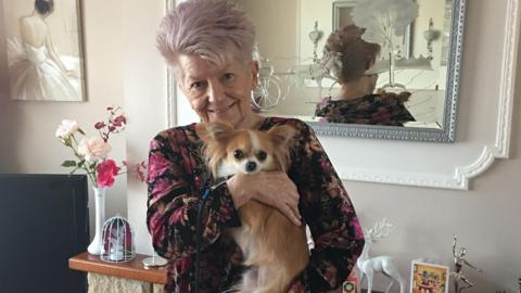 Madelin is smiling at the camera holding a small chihuahua. She's in a living room, there are small statues above a fire place and a small white vase with pink and purple roses standing in it. You can see Madelin's reflection in a mirror that is behind her.
