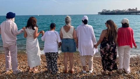 Seven people stand hand in hand on a pebble beach with their backs to the camera, looking out to sea towards a pier