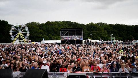 A crowd of hundreds of people stand at the front of the main stage at the park. A ferris wheel can be seen behind then, as well as a stand with the words Godiva Festival.