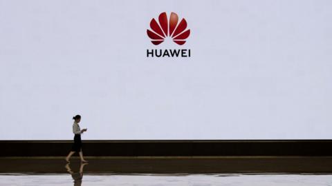 A member of Huawei's reception staff walks in front of a large screen displaying the logo