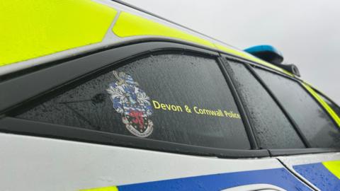 Exterior of a Devon and Cornwall Police car. The police logo is displayed on the car's window. Rain droplets are running down. 