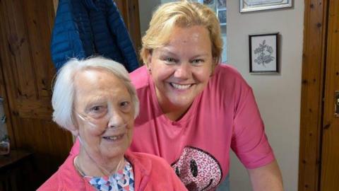 Denise Baxter, a woman with blond hair and wearing a pink T-shirt, smiles for the camera while standing next to Margo Baxter, a woman with white hair and a pink top. 