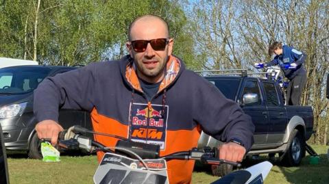 Wojtek Rozmus wearing a blue and orange jumper, standing next to a motocross bike, with the number 444 on it, and holding its handlebars.