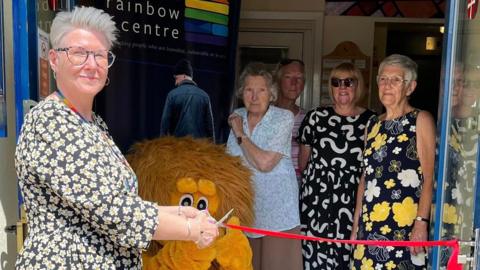 Jo Laking is pictured in a black dress with white flowers cutting a red ribbon with scissors for the opening of the new food bank as volunteers and a mascot look on