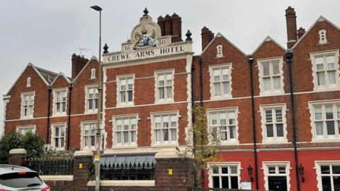 An image of Crewe Arms Hotel - a large 19th Century red brick building with a large crest at the top which says Crewe Arms Hotel.