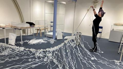 Claire Ward is in the process of installing her art called 'Catch of the Day'. It is a long string of plastic milk bottles which has been cut into long ribbons of plastic.