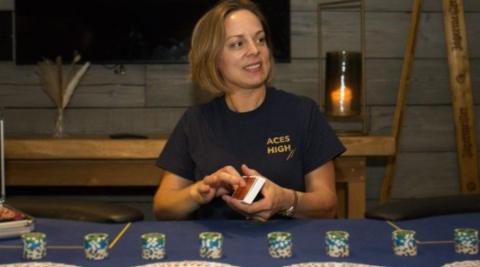 A woman sits at a table holding a pack of cards with stacks of betting chips laid out before her. She has shoulder length brown hair and is wearing a navy blue T-shirt. She is looking to her left at something that is out of shot, behind her is a wooden table with a lit candle on top of it.