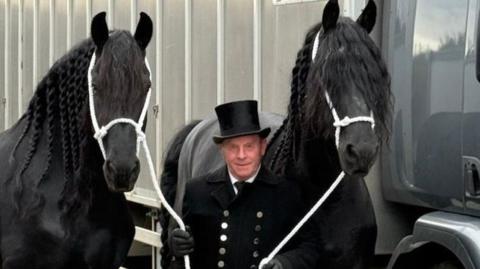 Modris Kesans with two black horses either side of him. He has a black jacket on with silver buttons and a black top hat. He holds the horses with white rope.