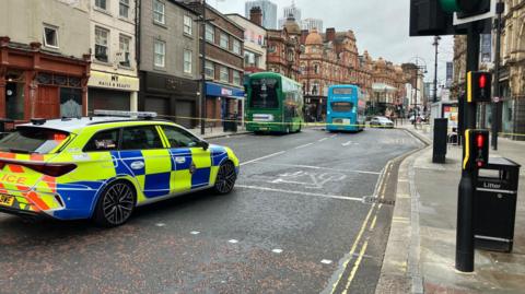 A road through a city centre with shops on either side. Part of the road has been cordoned off with yellow police tape. In the cordon there are two stationary double decker buses. Parked outside of the cordon is a police car. 