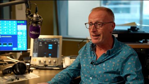 Man sat in a radio studio in front of computers and a microphone, he has short ginger hair and glasses and a blue shirt with patterns on it.