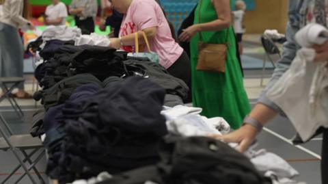 A table is loaded with folded uniform, which is being looked at by various people standing at the side of it.