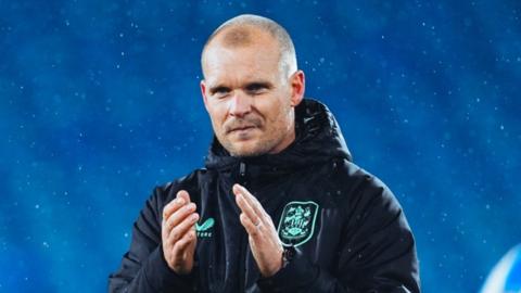 Liam Manning has a small smile on his face and claps his hands in front of his chest. He wears a navy rain jacket and stands in front of the blue seats of the Accu Stadium where it spits rain.
