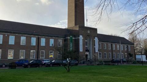 A view of the front facade of Salford Civic Centre with overhanging trees and a sunny sky in the background.