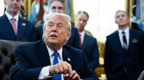 Donald Trump pictured sat behind his desk in the Oval Office. He wears a black suit jacket, white shirt, blue tie, and a pin of the US flag. Behind him stand four male officials in suits.